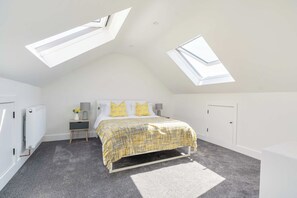 A bedroom with a sloped ceiling, skylights, a bed with a patterned bedspread, bedside lamps, and a radiator. - Lantern House | Historic Wells | Sleeps 11 (Somerset)