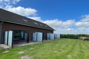 A building with a tiled roof, a grassy lawn, and a fenced area with a bench. - Holiday home with 2 bedrooms (Friedrichskoog-Spitze)