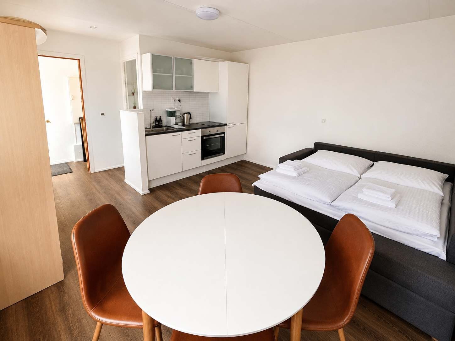 A compact hotel room with a kitchenette, a bed with white linens, and a round dining table with two chairs.
