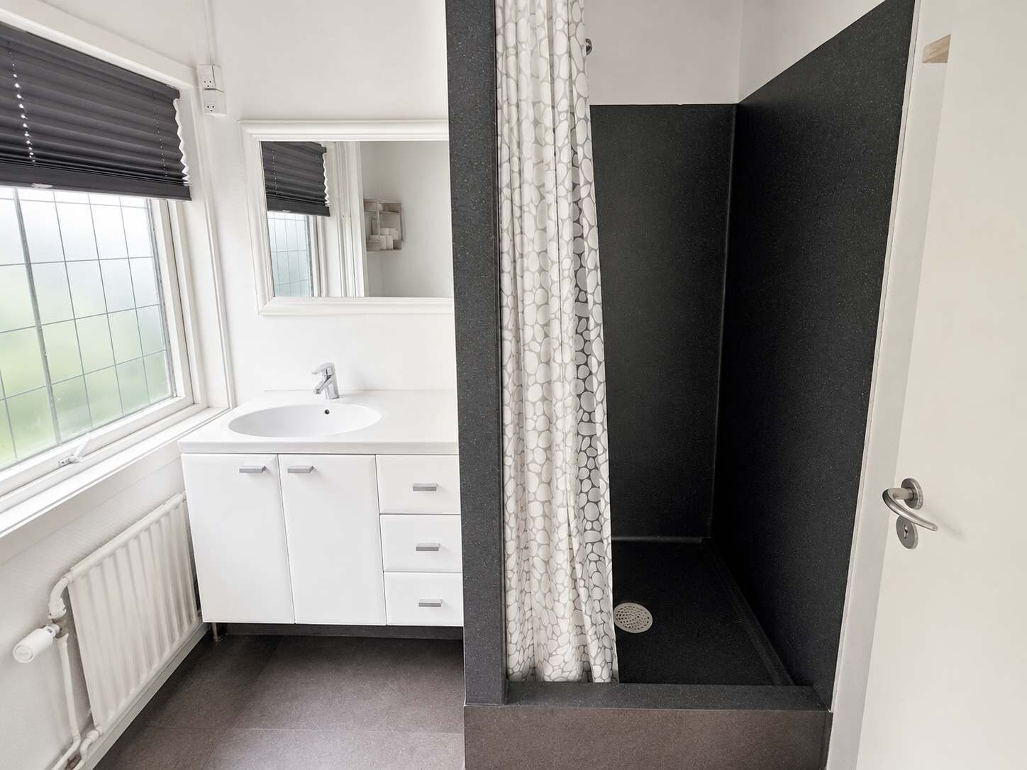 Et badeværelse med hvid vask, spejl, brusebad med mønstret gardin og en radiator.