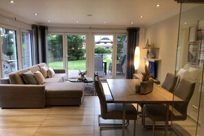 A modern living room with a sofa, dining table, and chairs. There is a glass door leading to a garden. - Holiday apartment with 1 bedroom (Norddeich)