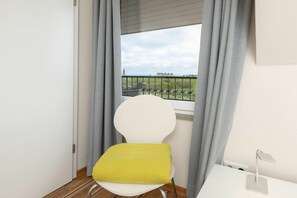 A room with a white chair, a small table, and a window with a view of a fence and buildings. - "Waddensee" at Uns to Hus (Juist)