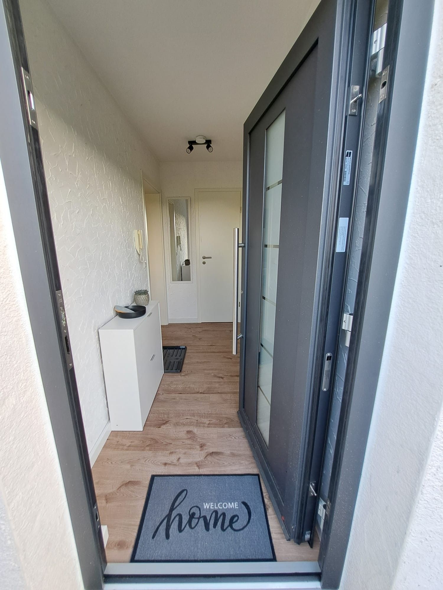 Ein schmaler Flur, der zu einer Tür mit Fußmatte 'Willkommen Zuhause' führt, mit einem kleinen weißen Schrank und einer Tür mit Milchglasverkleidung.