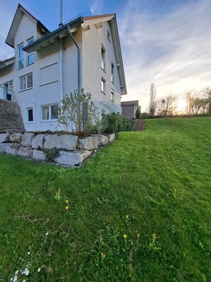Un edificio de dos pisos con base de piedra, techo plano y un pequeño jardín con césped y algunas plantas.