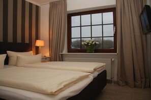 Ein Hotelzimmer mit Bett, Nachttisch mit Lampe, einem Fenster mit Vorhängen und einem an der Wand befestigten Fernseher.