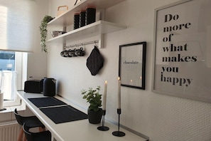 A modern kitchen with a white countertop, black mats, a potted plant, and a candle. There are shelves above with kitchen items and a framed motivational quote on the wall. - Holiday apartment with 1 bedroom (Lübeck)