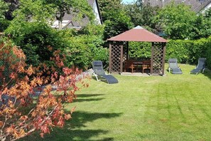 A gazebo with a red roof and a lattice structure, surrounded by a lawn with four lounge chairs. - Holiday apartment with 1 bedroom (Lübeck)