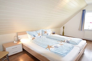 Ein Schlafzimmer mit schrägverdeckter Decke, einem Bett mit gestreifter Bettwäsche und Sternfischkissen, einem Nachttisch mit Lampe und einem Fenster mit Vorhängen.