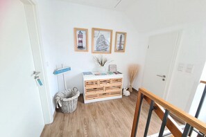 Ein Zimmer mit Holzboden, einer weißen Tür, einer weißen Wand mit drei gerahmten Bildern, einem weißen Schrank mit Schubladen, einem Korbwannen-Korb und einer kleinen weißen Figur.