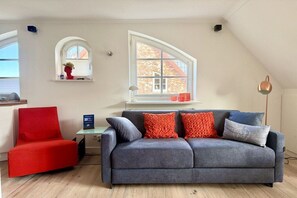 Ein modernes Wohnzimmer mit einem grauen Sofa, einem roten Sessel und einem großen Fenster mit Blick auf ein Backsteinhaus.