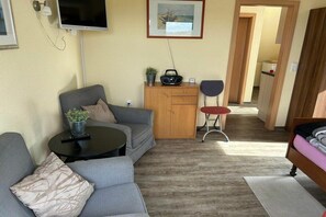 A compact living space with a sofa, a small table, a TV mounted on the wall, and a kitchenette area. - Holiday apartment in Juist with swimming pool (Juist)