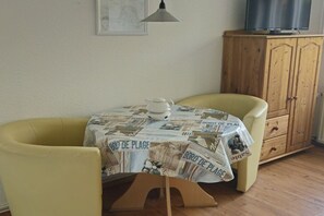 Ein Essbereich mit einem Tisch, der mit einer Tischdecke mit Zeitungsdruck bedeckt ist, zwei gelben Stühlen, einer Holzschrank und einem Fernseher auf dem Schrank.