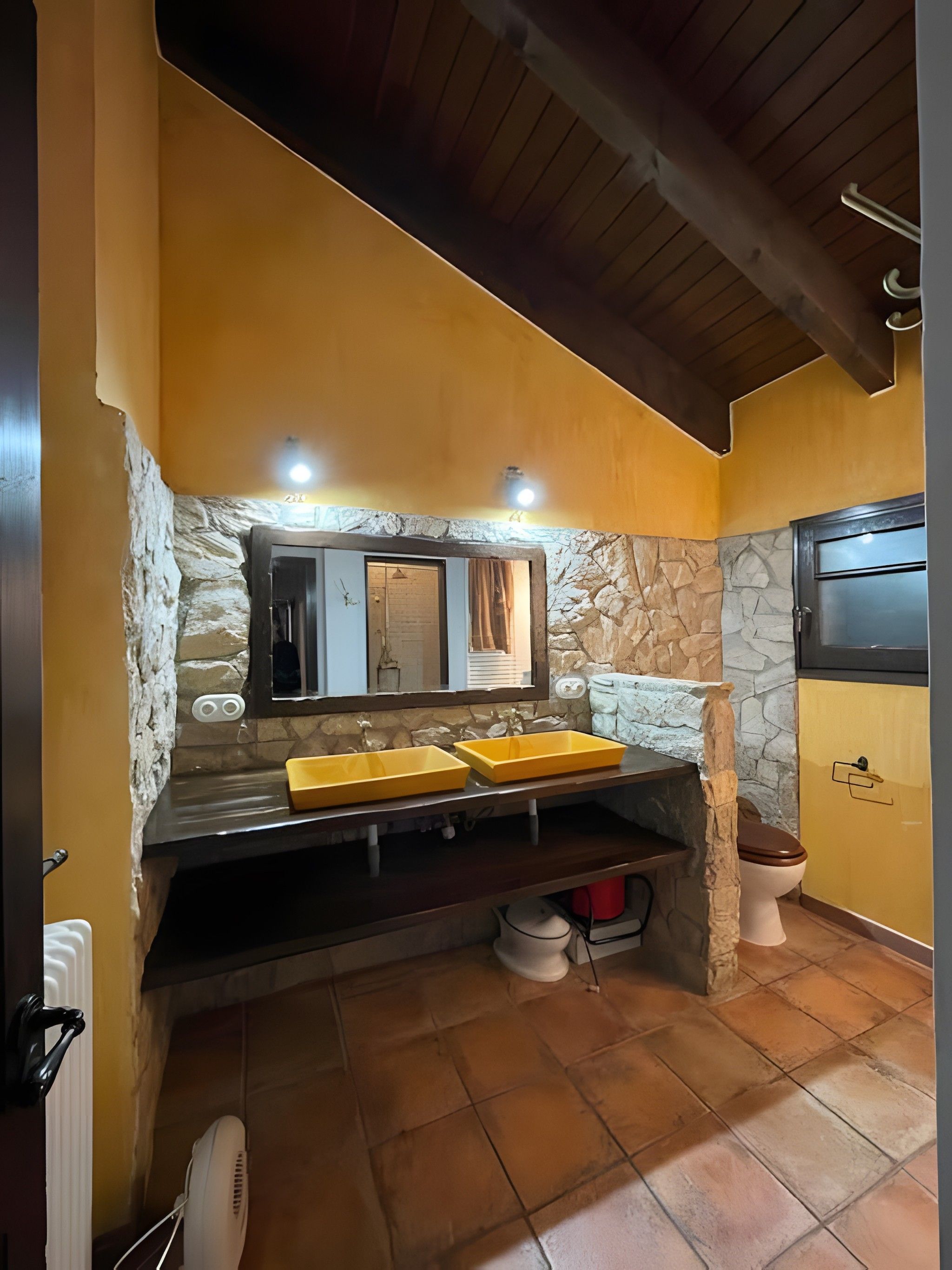 A bathroom with a stone wall, a yellow sink, a mirror, and a toilet.
