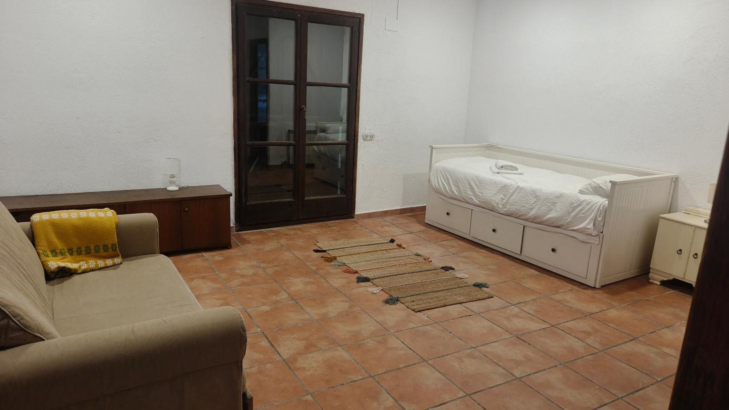 Una habitación pequeña y ordenada con una cama, un sofá, un mueble pequeño y una puerta que da a otra habitación.