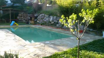 Una piscina con borde de piedra, un tobogán y un pequeño árbol en primer plano.