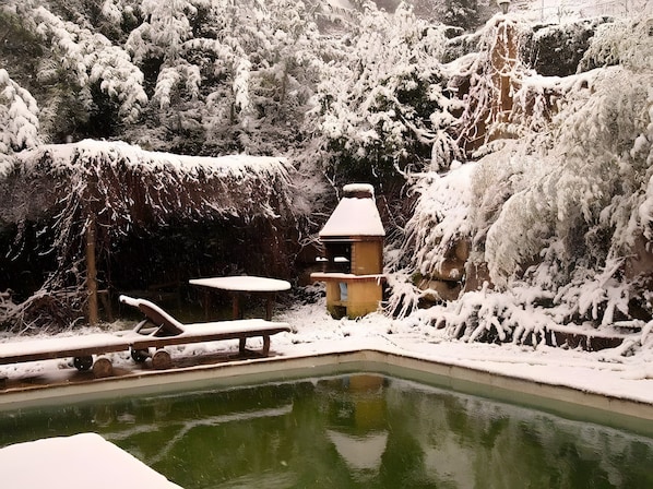 Un espacio exterior cubierto de nieve con piscina, un banco de madera y una pequeña construcción.
