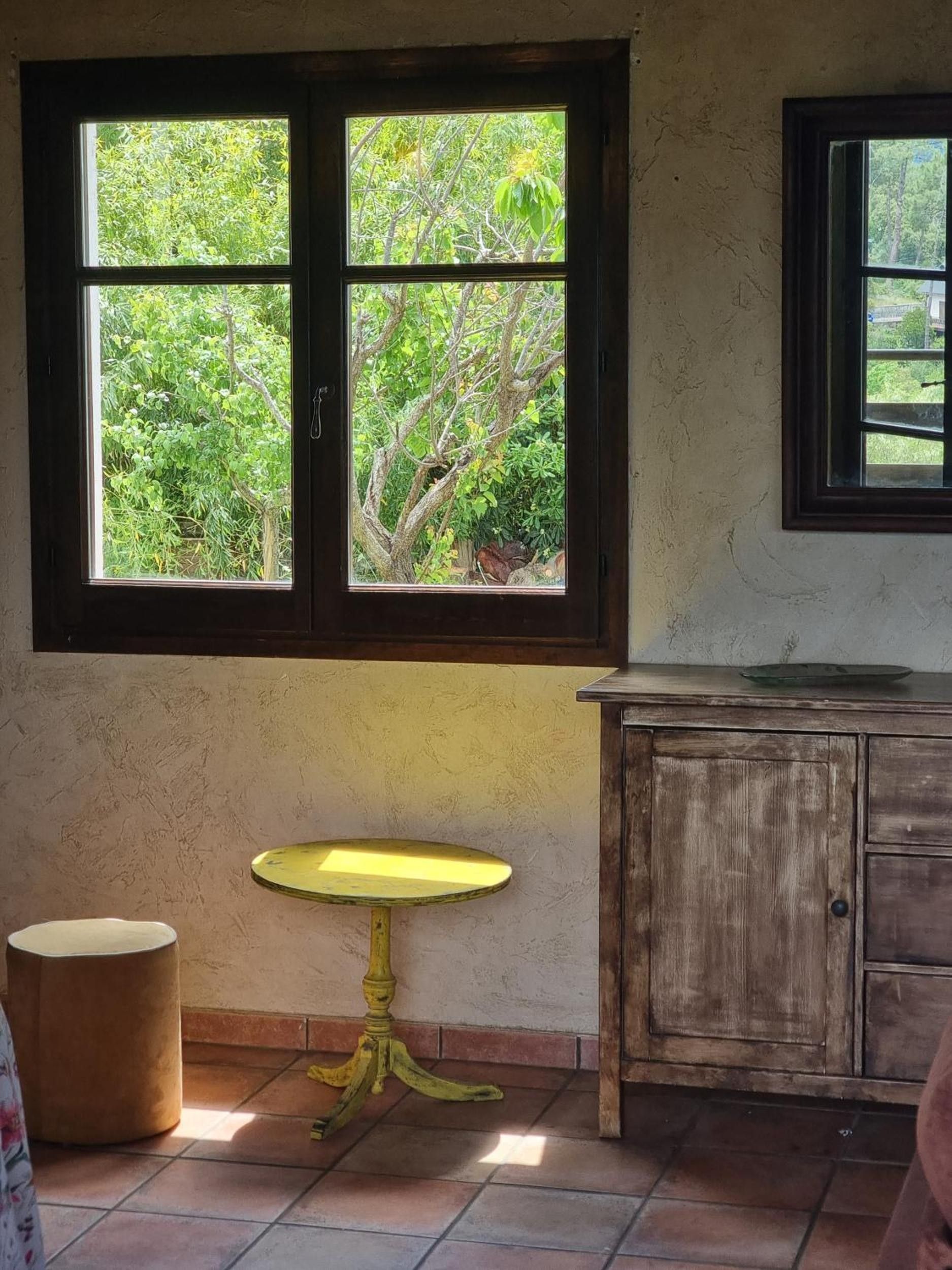 Quarto com janela para a área verde, uma pequena mesa amarela e um armário de madeira.