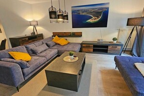 A modern living room with a sectional sofa, a coffee table, and a large painting of a coastal landscape. - Holiday apartment with 1 bedroom (Prerow)