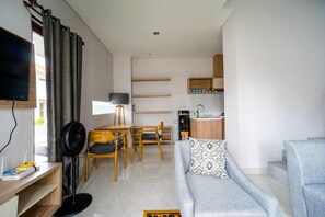 A modern living room with a grey sofa, a dining area with wooden furniture, a kitchen with wooden cabinets, and a flat-screen TV mounted on the wall.