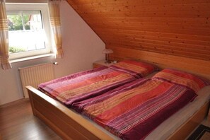 Ein Schlafzimmer mit schrägen Wänden, einem hölzernen Bett mit gestreifter Decke, einer Nachttischlampe und einem Fenster mit Vorhängen.