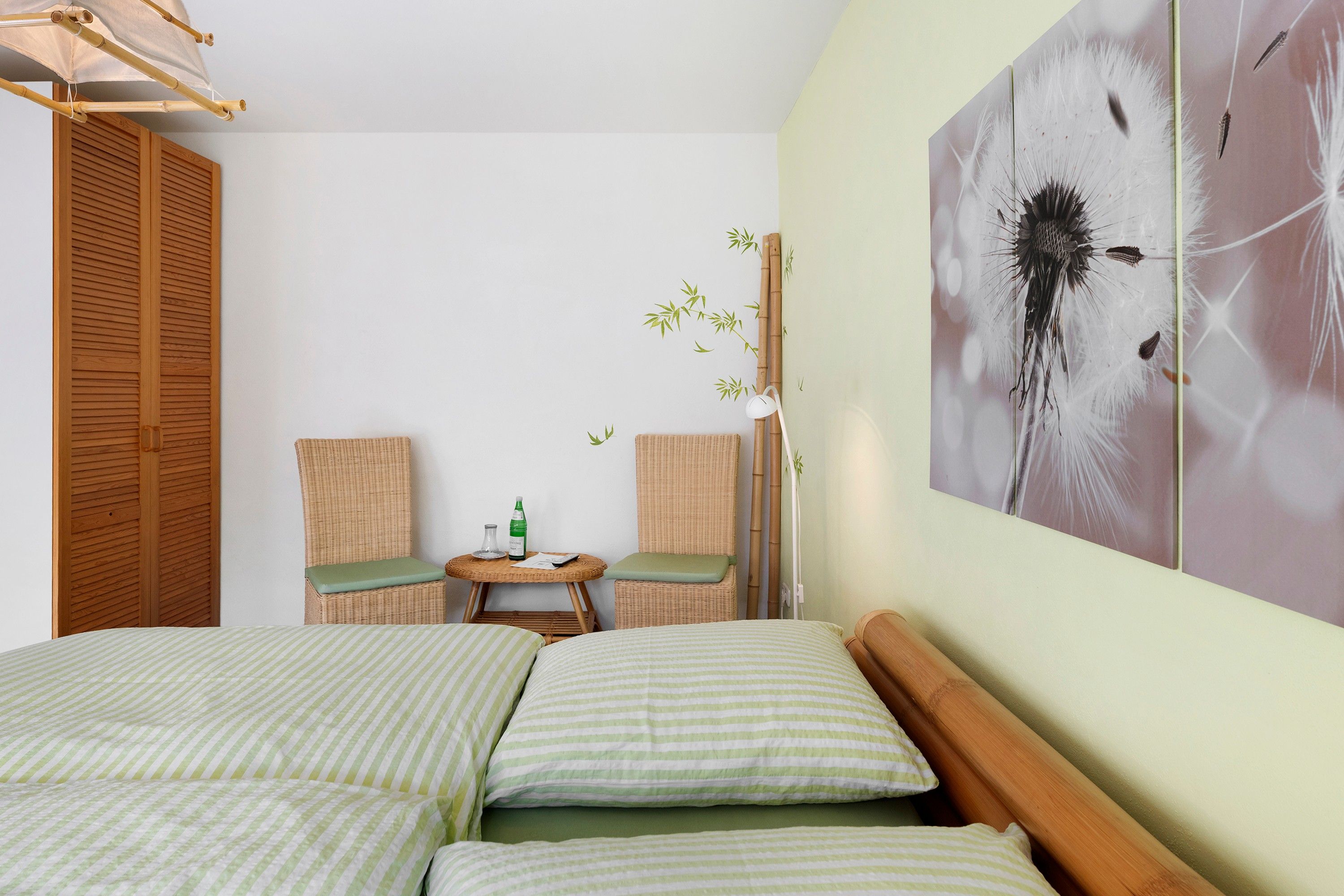 Una habitación con una cama, dos sillas, una mesita con una botella y un control remoto, y una pared decorada con una obra de arte de diente de león.