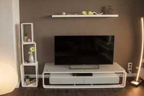A white entertainment unit with a flat-screen TV, shelves with decorative items, and a wall-mounted shelf above. - Holiday apartment with 2 bedrooms (Wilhelmshaven)
