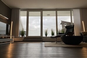 A modern living room with a large window, a black sofa, a flat-screen TV, and a potted plant. - Holiday apartment with 2 bedrooms (Wilhelmshaven)