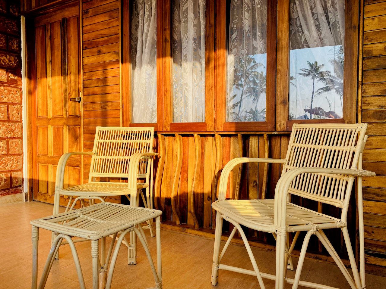 A room with two wicker chairs and a small table, wooden walls, and a window with curtains.