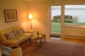 A living room with a sofa, a lamp, a coffee table, and a view of a body of water through a glass door. - Angelika von Lingelsheim (Ratzeburg)