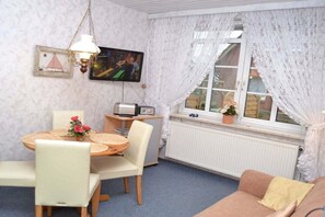 Ein Zimmer mit Esstisch, Stühlen, einem Fernseher, einem Fenster mit Spitzenvorhängen und einer Heizung.
