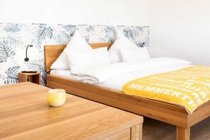 Ein ordentlich bezogenes Bett mit weißer Bettwäsche und einer gelben Decke mit Text, ein Nachttisch mit Lampe sowie ein Holztisch mit einer Kerze.