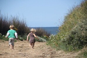Twee personen lopen over een zandpad, geflankeerd door begroeiing, met een waterlichaam op de achtergrond.