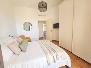A bedroom with a bed, pillows, a bedside lamp, a mirror, a TV, and a door leading to another room. - Click Sardegna Villa SeaSky – Sea View with Garde (Alghero)