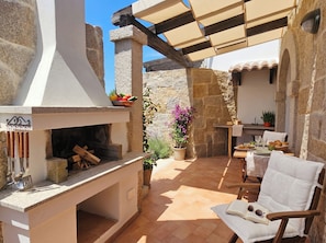 An outdoor patio with a stone fireplace, a table set for dining, and a chair with a book and sunglasses. - Click Sardegna Villa SeaSky – Sea View with Garde (Alghero)