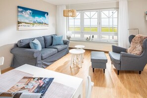 A living room with a grey sofa, a grey armchair, a white table, and a window with a view of the beach. - Holiday apartment Juister Gezeiten (Juist)