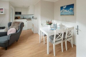 A modern living room with a grey armchair, a white dining table with four chairs, and a kitchen in the background. - Holiday apartment Juister Gezeiten (Juist)