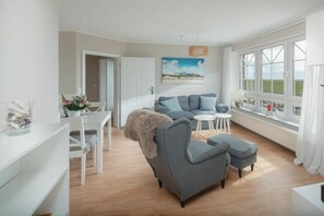 A modern living room with a grey sofa, a small round table, and a white coffee table. There is a window with a view of the outdoors. - Holiday apartment Juister Gezeiten (Juist)