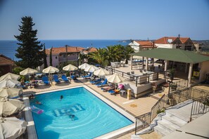 Ein Poolbereich mit Liegestühlen und Sonnenschirmen, freie Sicht auf das Meer und ein Gebäude mit rotem Ziegeldach.