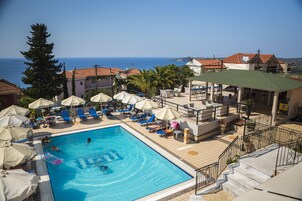 Zona de piscina con sillones de descanso y sombrillas, vista despejada del mar, y un edificio con tejado de tejas rojas.