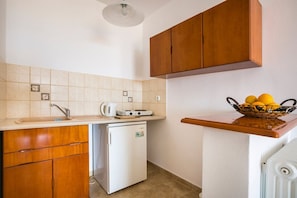 A compact kitchen with a sink, stove, and refrigerator, wooden cabinets, and a fruit basket on the countertop. - Saoulas Studios (Kefalonia)
