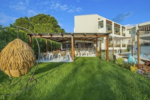 A modern outdoor dining area with a pergola, a hanging chair, and a poolside seating area. - Mirablue Deluxe Residences (Kassandra)