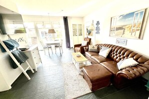 A living room with a brown leather sofa, a coffee table, and a television. There is a dining area with a table and chairs. - Holiday apartment with 1 bedroom (Norddeich)