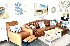 A living room with a brown leather sofa, a wooden coffee table, and a white armchair with an anchor pillow. - Holiday apartment with 1 bedroom (Norddeich)