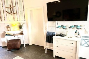 A room with a flat-screen TV mounted on the wall, a desk with a lamp, a chair, and a small stool. - Holiday apartment with 1 bedroom (Norddeich)