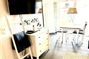 A room with a flat-screen TV mounted on the wall, a white dresser with drawers, a small table with a chair, and a window with a wicker lamp. - Holiday apartment with 1 bedroom (Norddeich)