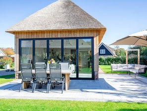 Ein modernes Haus mit einem Strohdach, großen Glasfenstern und einer Terrasse mit Gartenmöbeln.