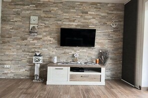 A living room with a stone wall, a flat-screen TV mounted above a white entertainment unit, and a wooden floor. - Holiday home in Aurich with swimming pool (Aurich)
