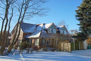 Ein zweistöckiges Haus mit schneebedecktem Dach, umgeben von kahlen Bäumen und einem klaren Himmel.