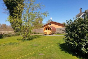 Eine Holzhütte mit gewölbtem Dach, vermutlich eine Sauna oder ein Whirlpool, in einer grünen Umgebung mit Holzzaun und einem Haus im Hintergrund.