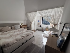 A bedroom with a large bed, bedside tables, a desk, and a window with sheer curtains. - Bright Top Floor Flat with Balcony in Purley (London)